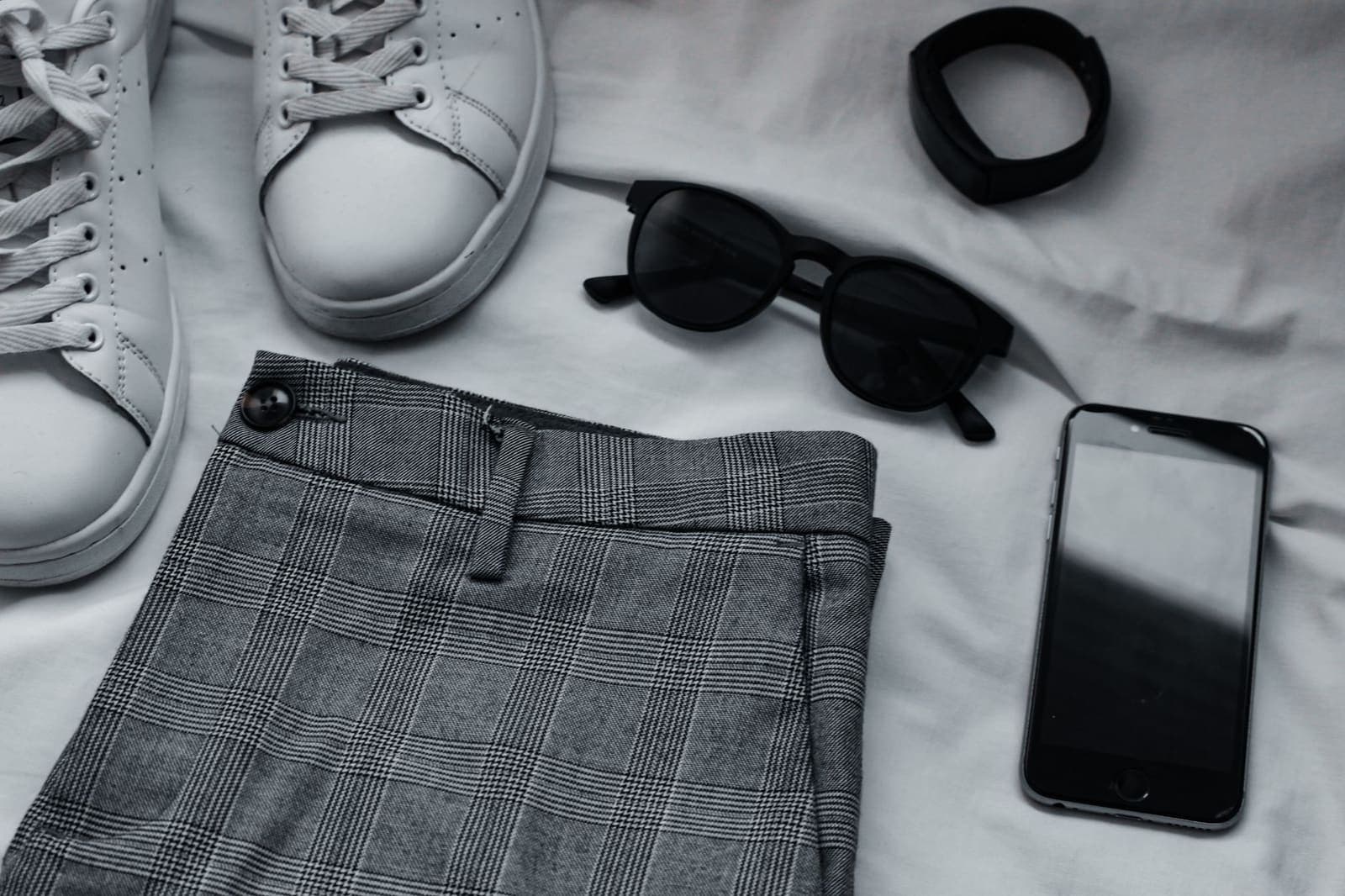 Sneaker, Sonnenbrille, Hose und Handy nebeneinander auf hellem Untergrund